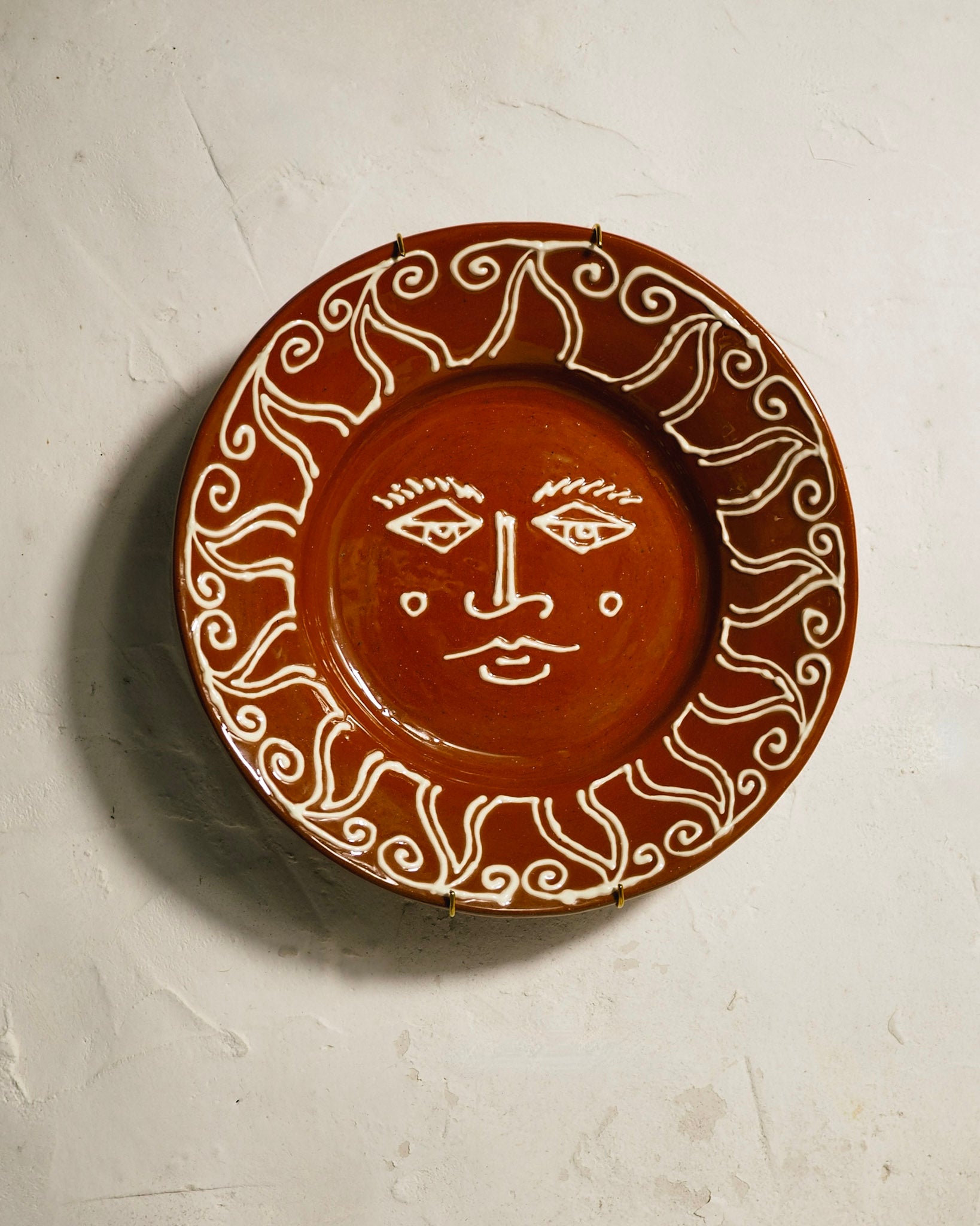 A round terracotta ceramic wall plate featuring a hand-painted white face in the center and swirling sunburst patterns around the rim, mounted on a textured off-white plaster wall.