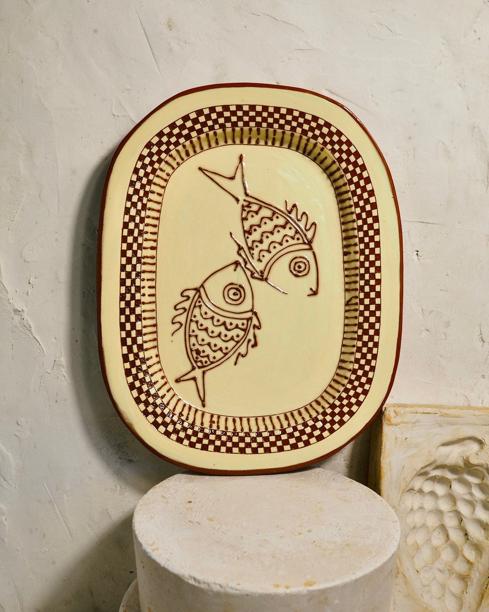 An oval ceramic serving platter with a cream-colored glaze, featuring two brown fish illustrations and a brown checkerboard border, displayed on a textured white wall above a stone pedestal.