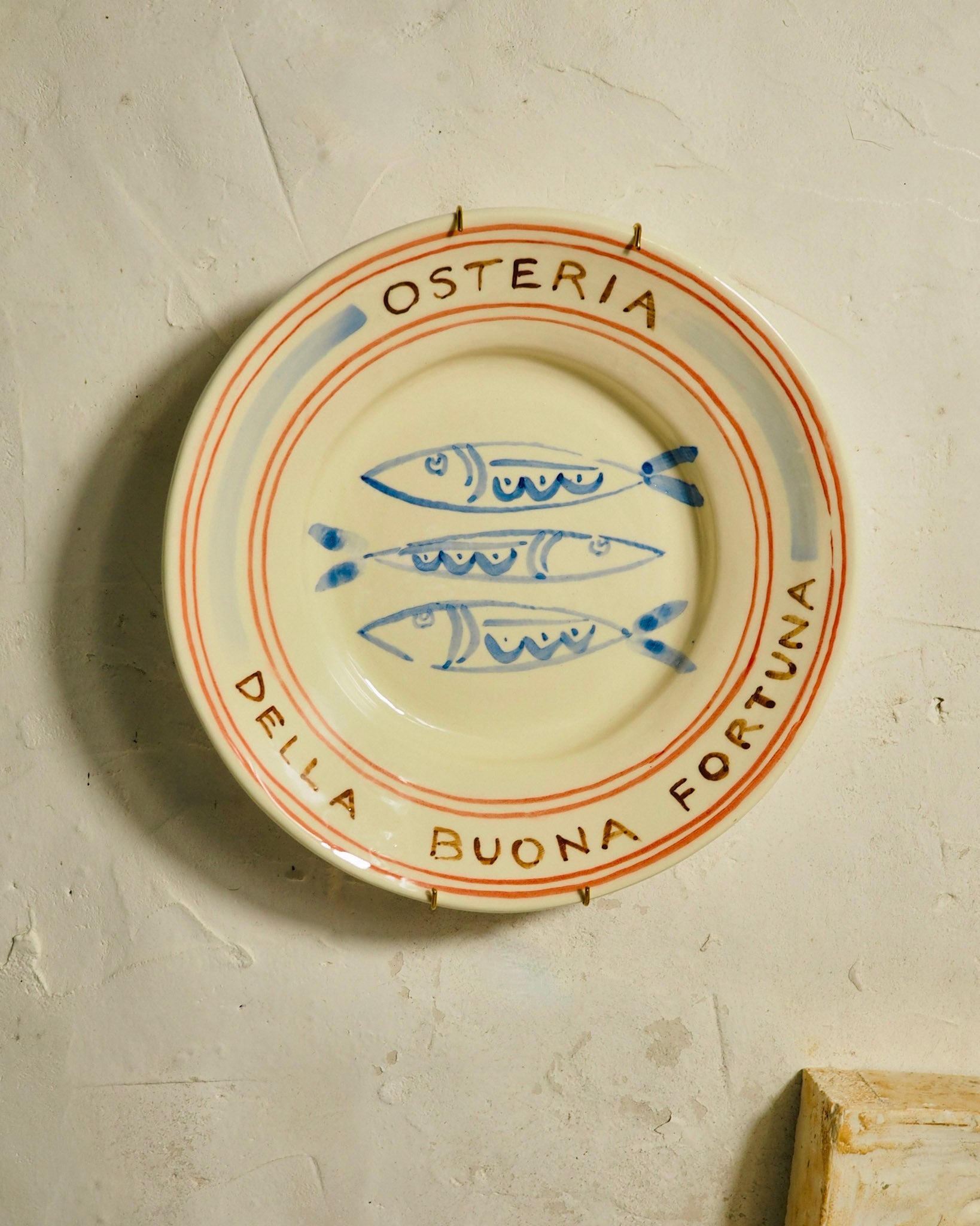 A round ceramic plate with a cream-colored glaze, featuring blue painted fish motifs in the center and orange-brown text reading 'OSTERIA DELLA BUONA FUORTUNA' around the rim, mounted on a textured plaster wall.