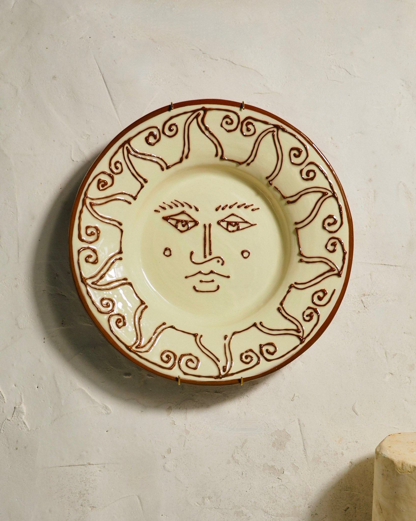 A round ceramic wall plate featuring a brown sun face design in the center and a matching sunburst border, mounted on a textured off-white plaster wall.