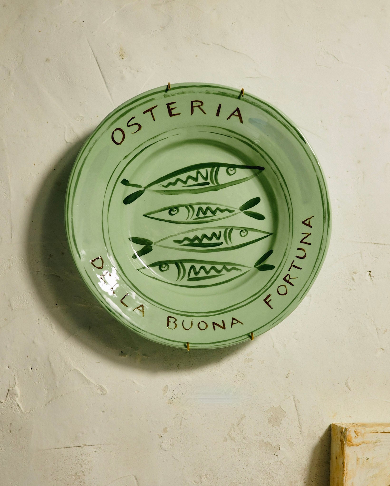 A round light green ceramic plate with hand-painted dark green fish illustrations and the text 'OSTERIA DELLA BUONA FORTUNA' on a textured off-white plaster wall.