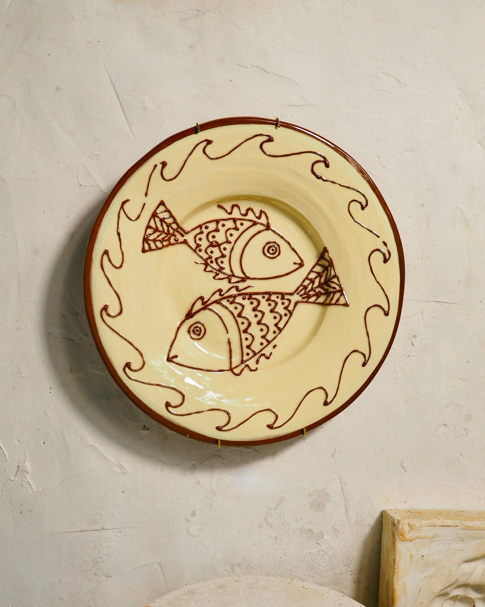 A round ceramic wall plate with a cream-colored glaze, decorated with two brown fish and wavy border lines, mounted on a textured off-white plaster wall.