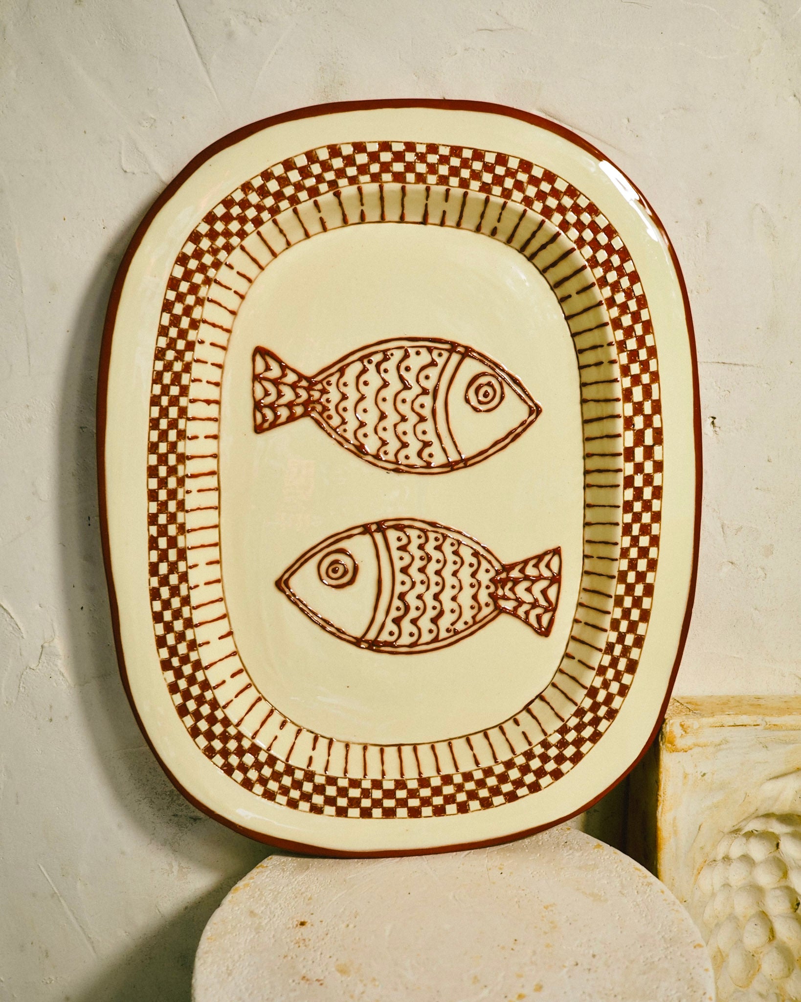 An oval ceramic serving platter with a cream-colored glaze and a brown hand-painted design featuring two fish and checkered borders, displayed on a textured light gray surface.