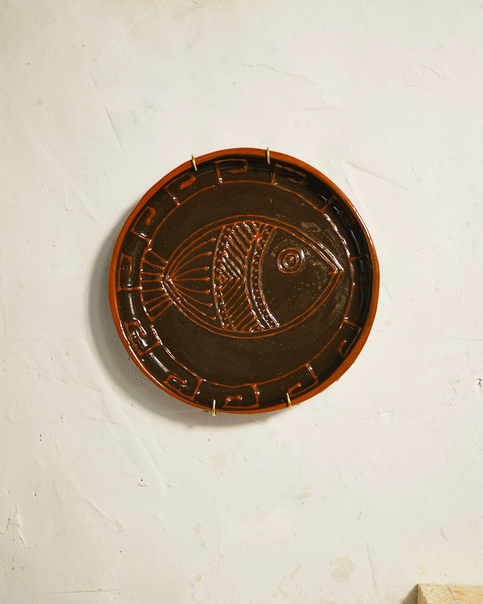 A round dark brown glazed ceramic plate featuring an embossed fish design in the center and geometric border patterns, mounted on a textured off-white wall.