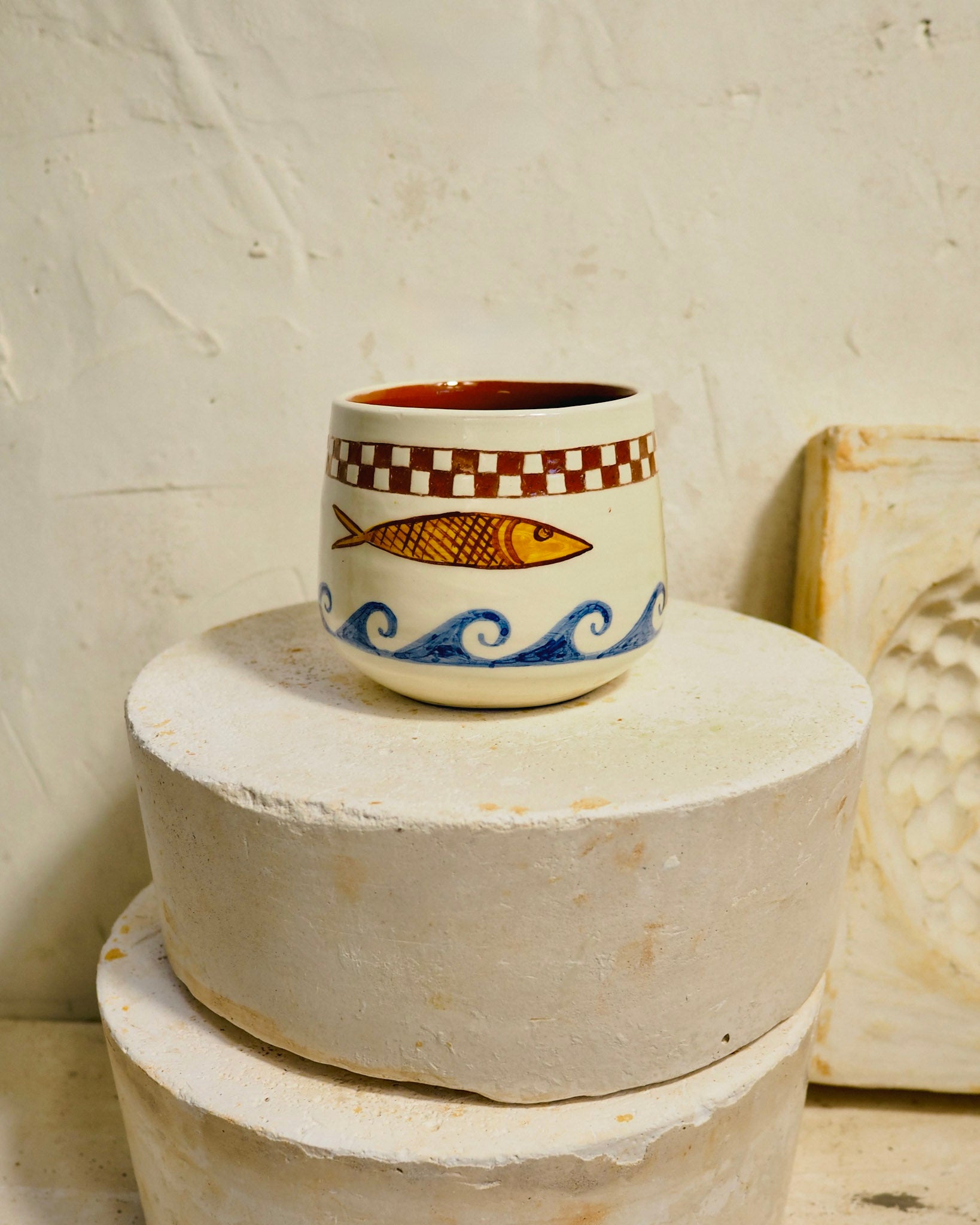 A small handmade ceramic cup with a fish and wave design, displayed on stacked round stone blocks against a textured cream wall.