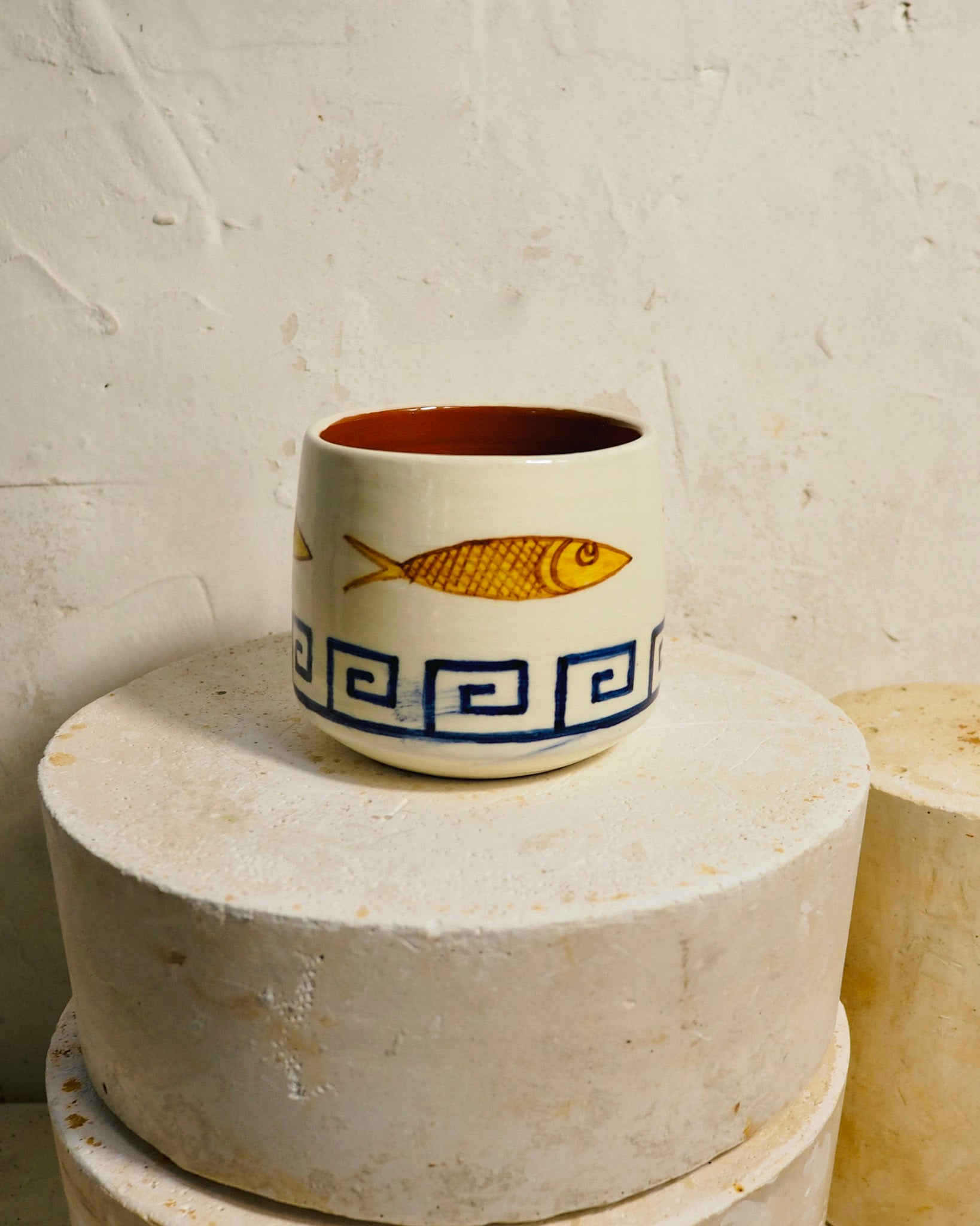 A small ceramic cup with a warm terracotta interior, decorated with a yellow fish and a blue Greek key pattern on a cream-colored exterior, displayed on a textured stone pedestal against a rustic plaster wall.