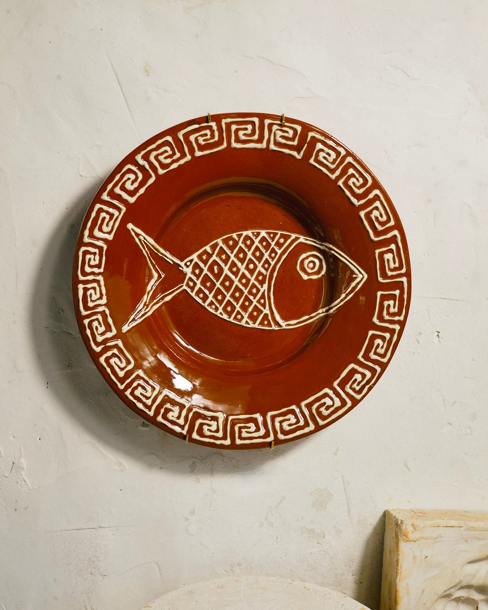 A round terracotta-colored ceramic plate with a white Greek key border and a white fish design in the center, displayed against a textured off-white wall.