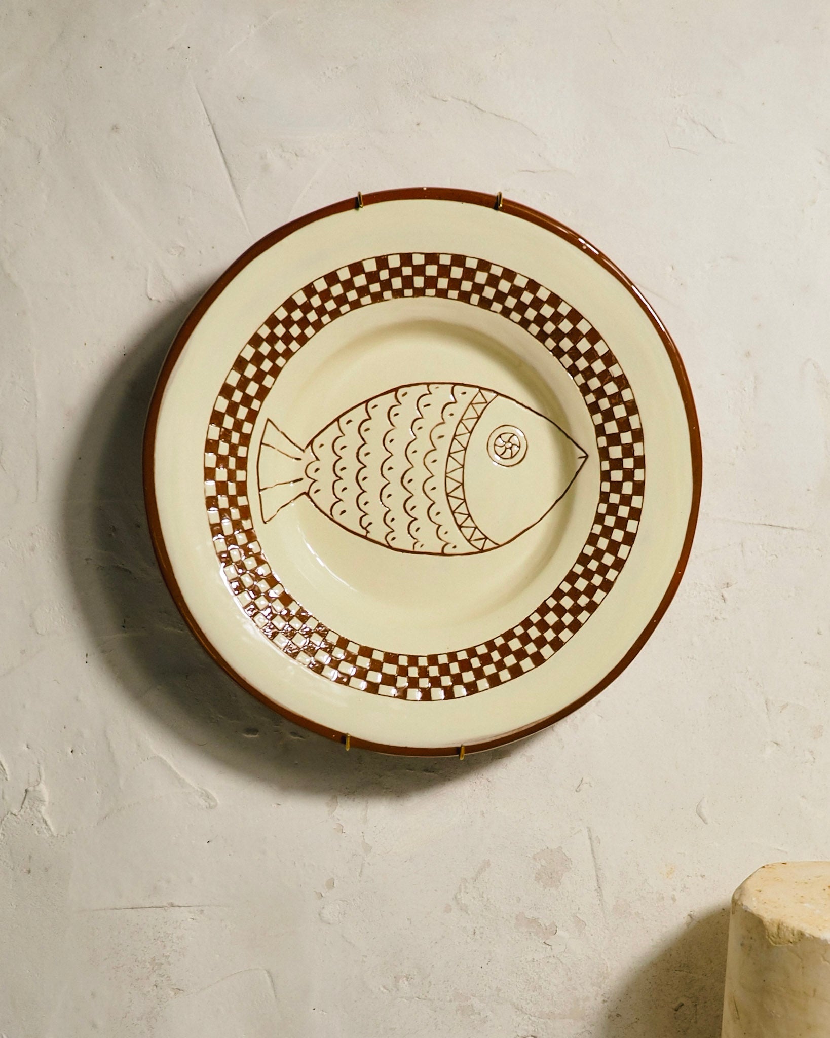 A round ceramic plate with a brown fish motif and checkered border, displayed against a textured off-white wall.